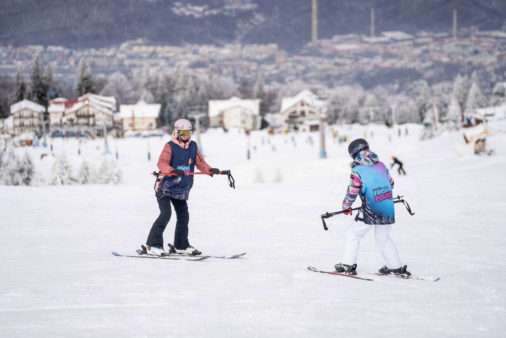 Two skiers glide on a snowy slope with a town and snow-covered trees in the background, during daytime winter scenery.