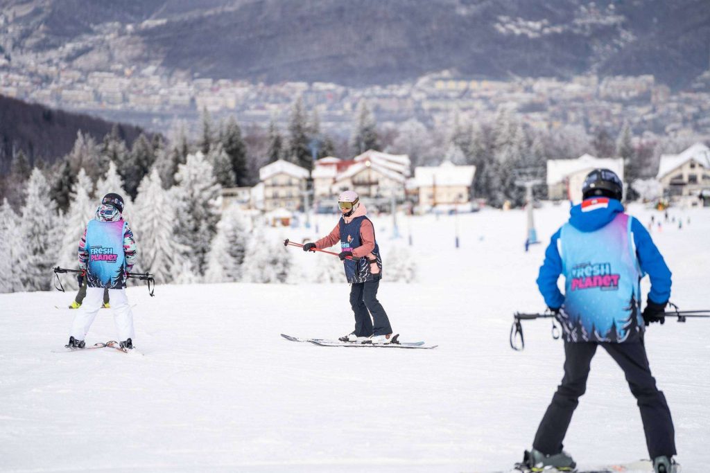 Group of skiers on a snowy slope with a town and frosted trees in the background, wearing blue jackets that say Fresh Planet on the back.