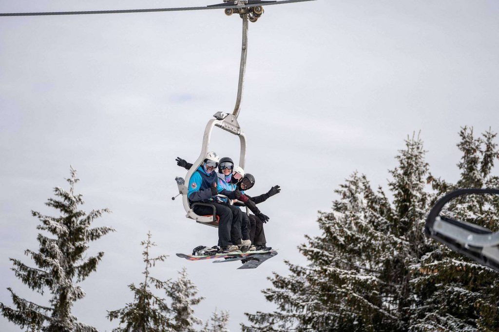 Three snowboarders wearing helmets ride a chairlift over a snow-covered landscape, trees below.