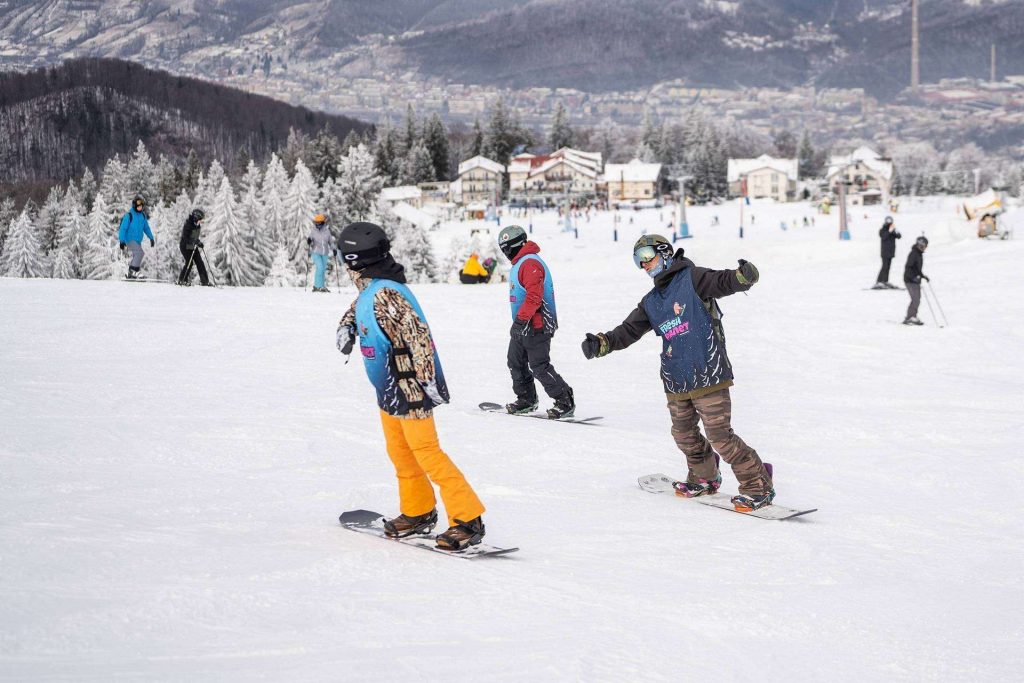 Snowboarders and skiers glide down a snowy slope with snow-covered trees and a village in the distance.