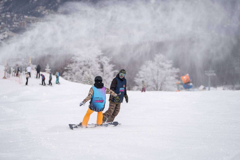 Two snowboarders holding hands on a snowy slope, bright jackets visible in the foreground.