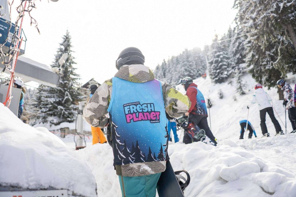 Person wearing a blue vest with Fresh Planet logo stands in a snowy ski area, facing away as others prepare to ski nearby.