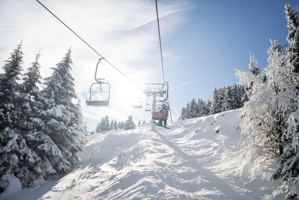 Snowy mountain scene with a chairlift carrying skiers above a slope lined by snow-covered evergreen trees.