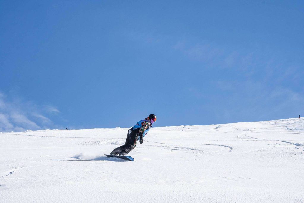 Snowboarder in colorful gear carving down a snowy slope under a clear blue sky.