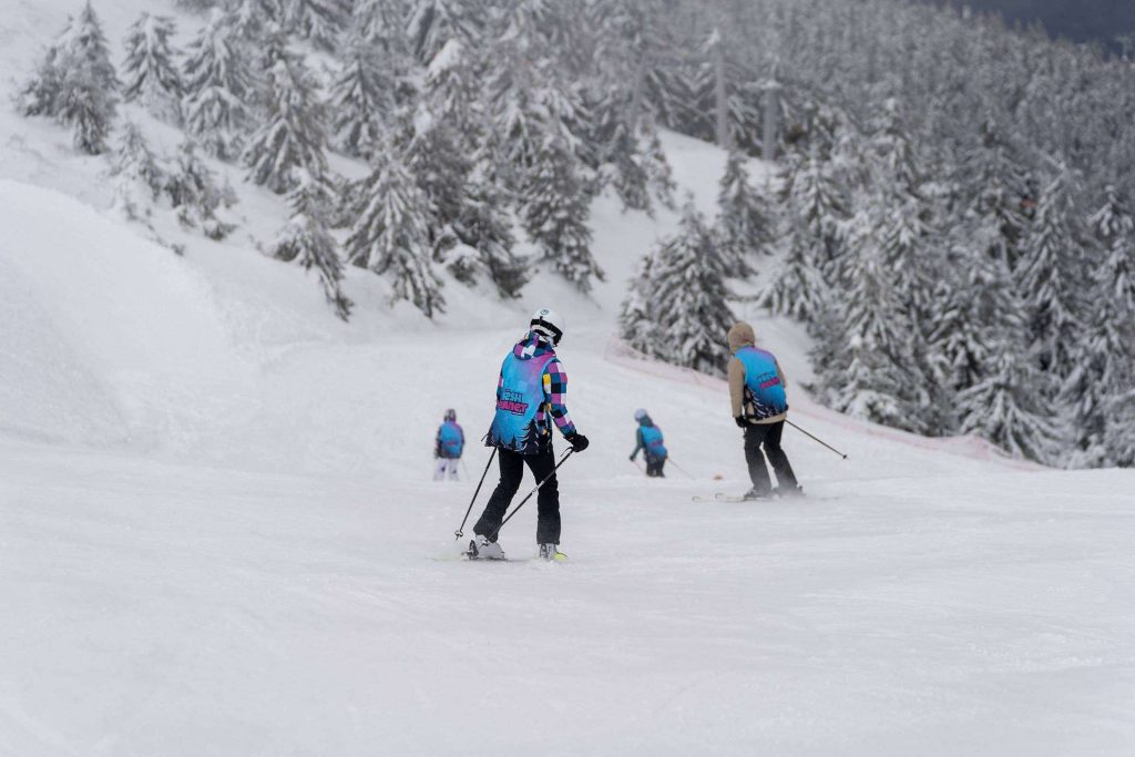 Skiers in bright jackets descend a snowy slope with snow-covered evergreen trees in the background.