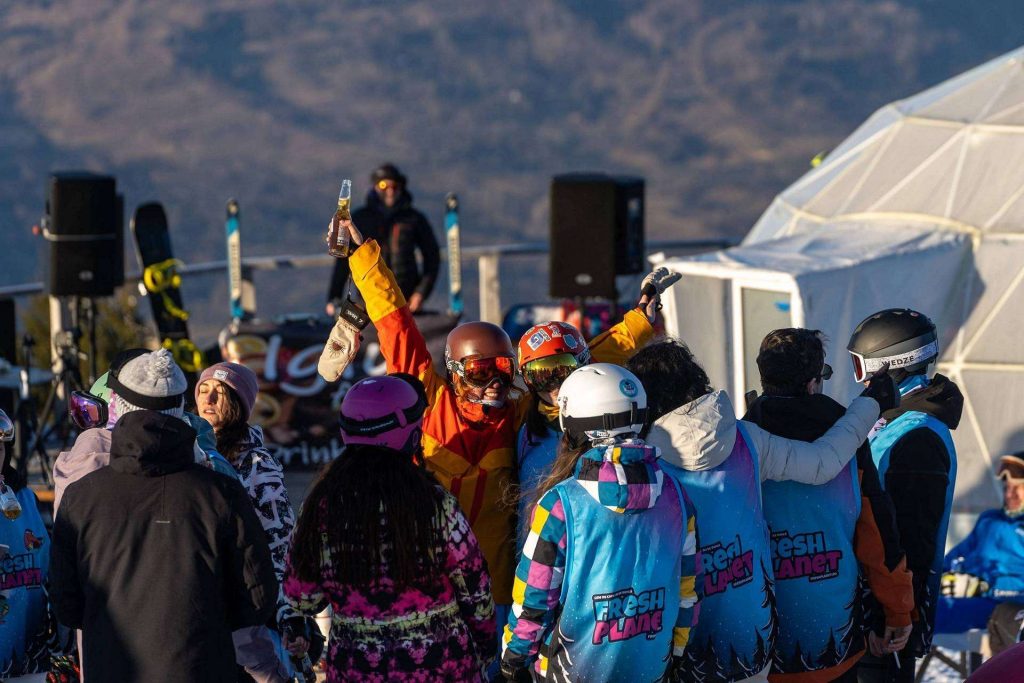 Group of skiers and snowboarders celebrate, raising bottles in a outdoor snow event with tents in the background.