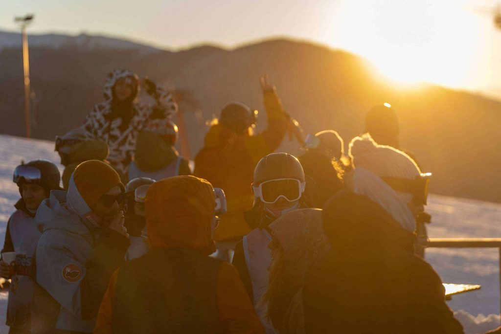 Group of skiers and snowboarders in jackets and goggles gathered on a snowy slope at sunset.
