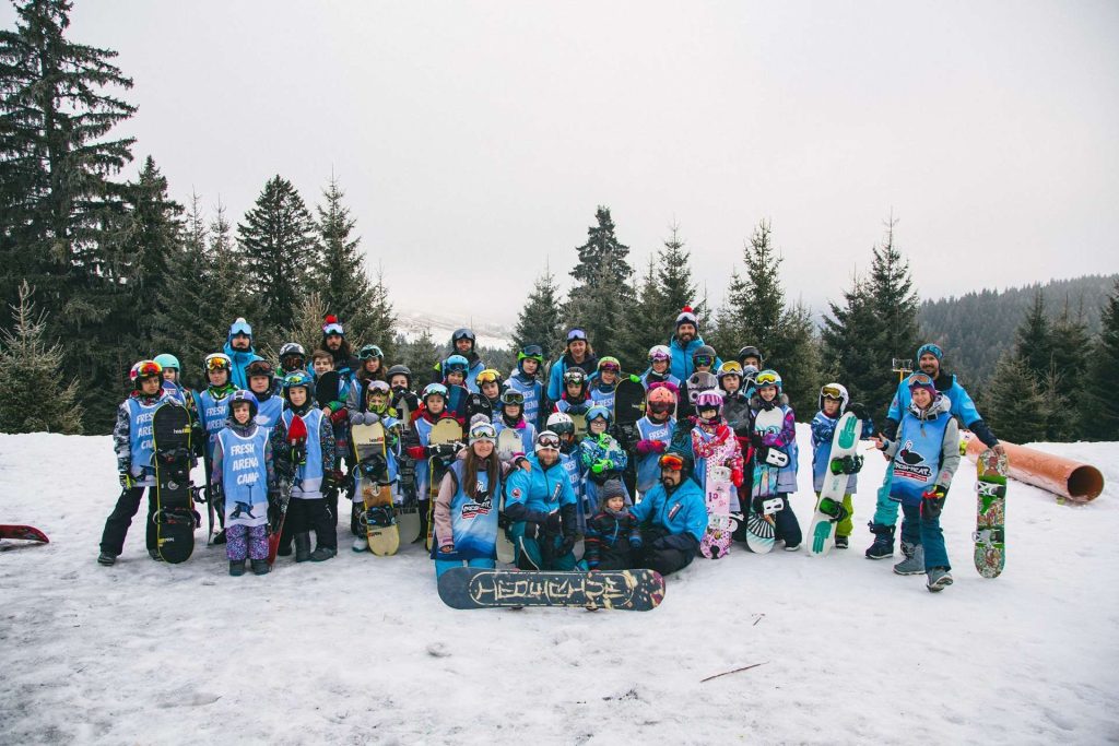 Group of children and adults in winter gear posing with snowboards on a snowy slope, forest trees in the background.