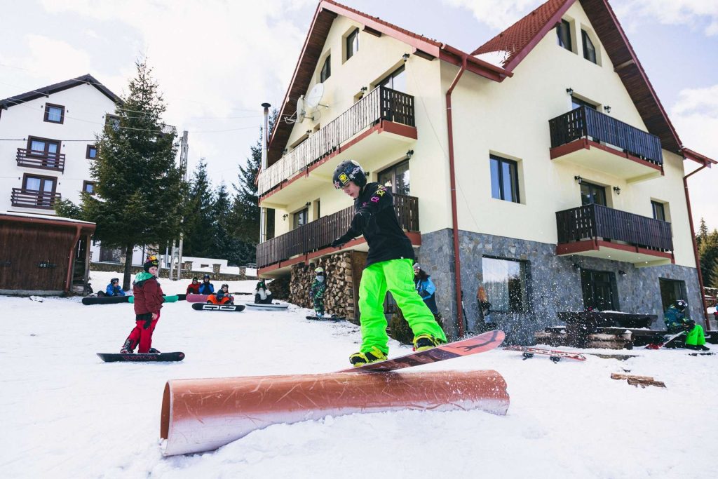 Snowboarder in neon green pants jumps over a snow obstacle in front of a chalet-style building; other riders watch nearby.