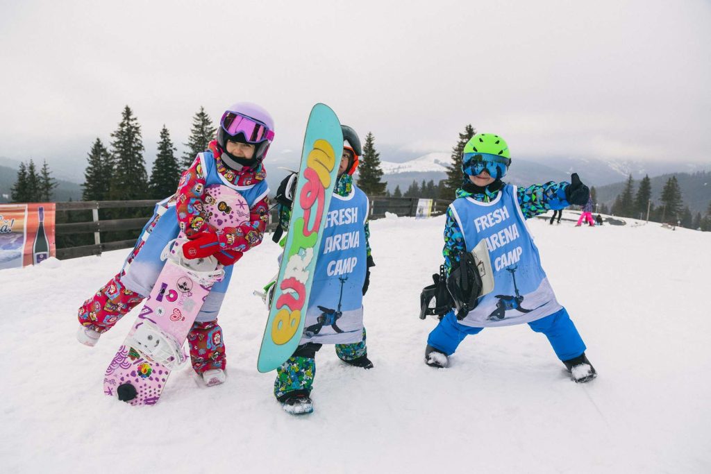 Three kids in colorful snow gear and helmets pose with their snowboards on a snowy slope, wearing blue 'Fresh Arena Camp' vests.