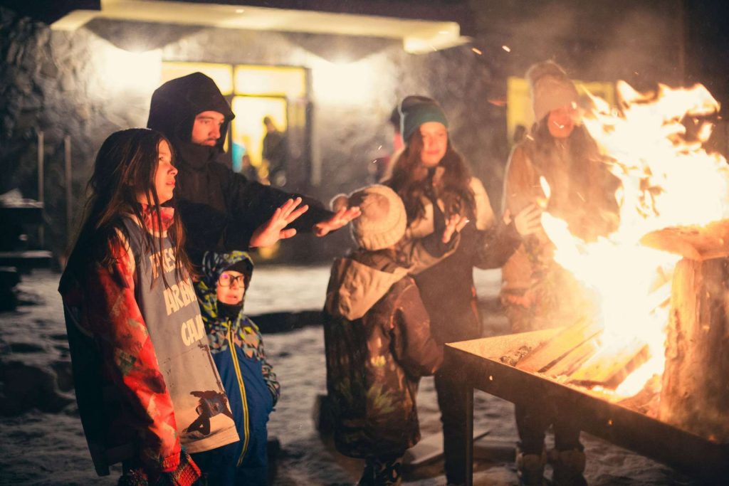 Group of children and adults gathered around a large bonfire at night, reaching toward the flames.