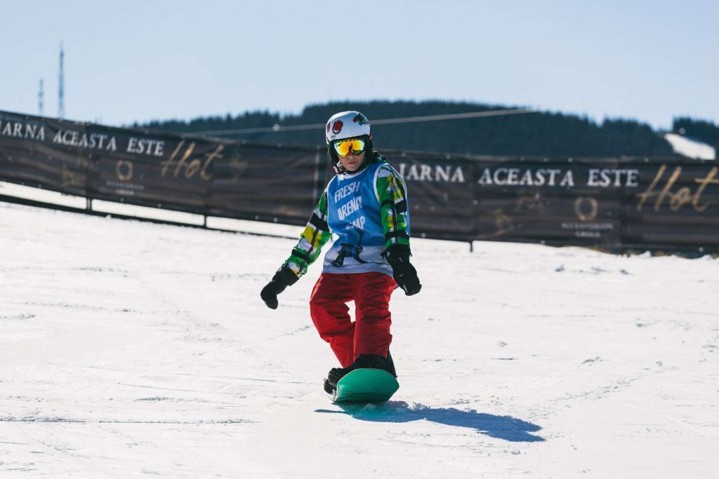 Young snowboarder in colorful jacket and red pants gliding on a snowy slope with a dark banners in the background.