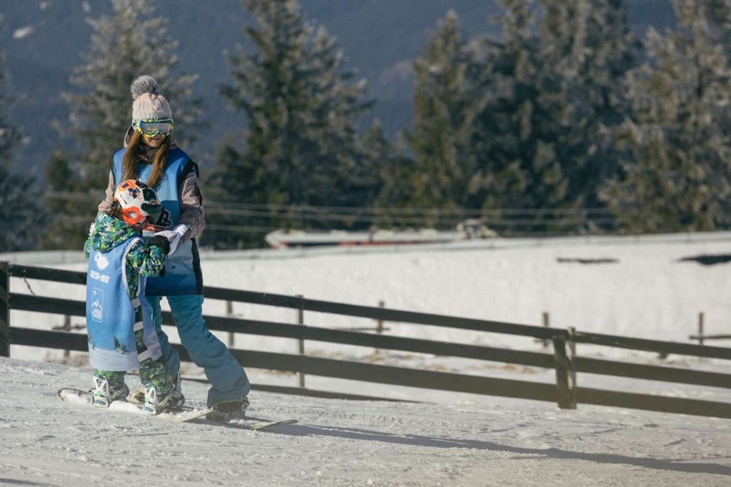 A skier in blue gear helps a child in a green patterned snowsuit with a helmet on a snowy slope beside a wooden railing, mountain trees in the background.