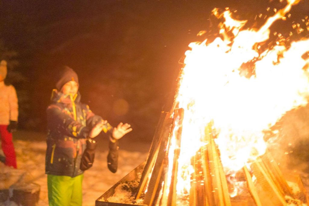People in winter jackets standing around a tall campfire, arms raised toward the flames at night.