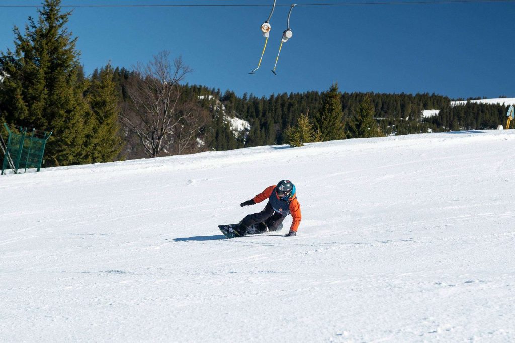 Snowboarder in an orange jacket carves down a sunlit snowy slope with pine trees in the background, under a blue sky.