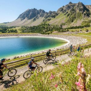 Group of mountain bikers riding along a dirt trail beside a turquoise alpine lake with rocky shore and rugged mountains in the background.