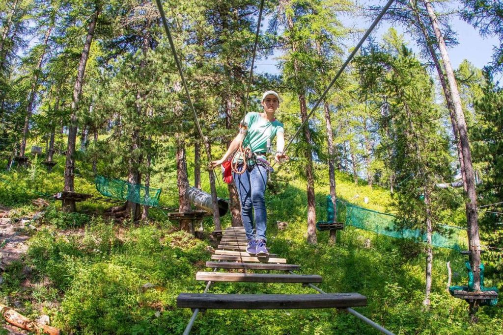 Person walking a suspended wooden bridge in a forest adventure park, wearing a helmet and safety harness.