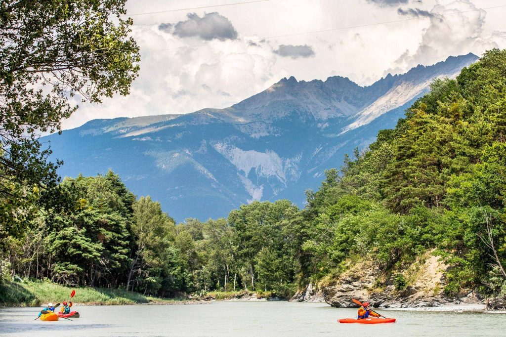 Two kayakers paddle on a calm river, surrounded by green forest and distant snow-capped mountains.