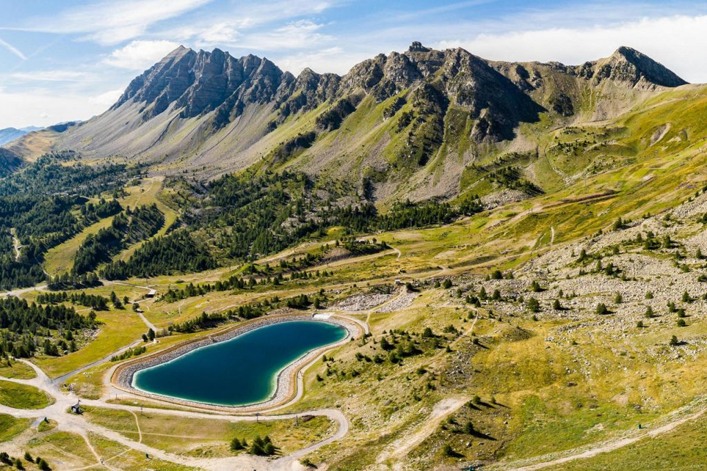 Heart-shaped turquoise lake nestled in a grassy alpine valley with winding trails and rugged mountains in the background.