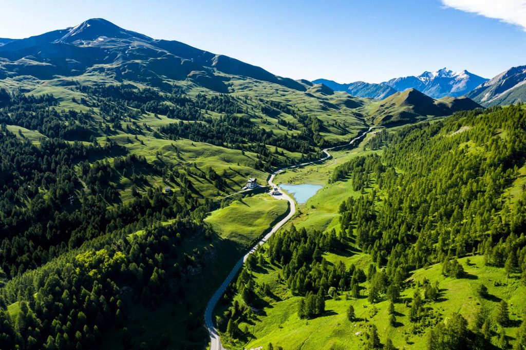 A winding road runs through a green Alpine valley with forests, a small lake, and distant snow-capped mountains.