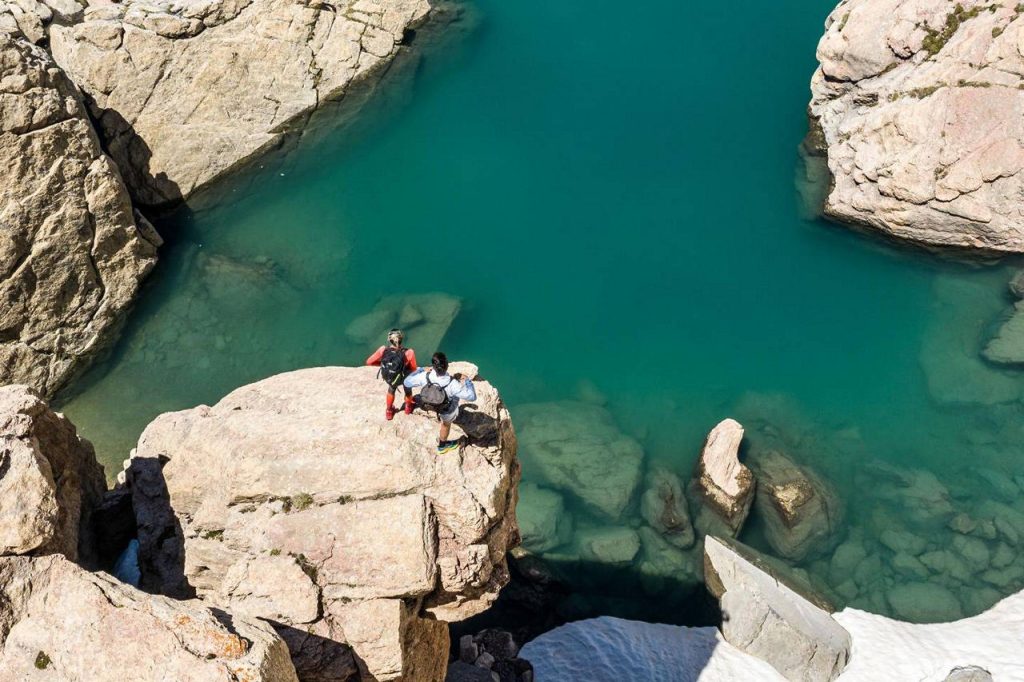 Two hikers in gear stand on a rocky ledge overlooking a turquoise alpine lake surrounded by canyon walls.