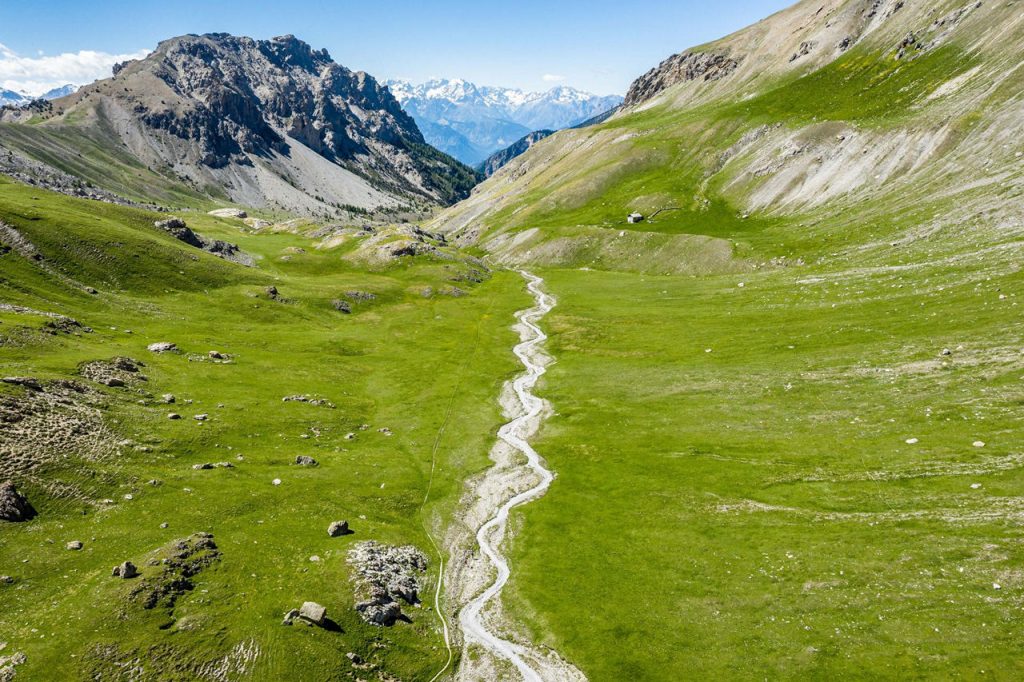 Alpine valley with a winding dry creek through green meadows and distant snow-capped peaks, a small hut on the hillside