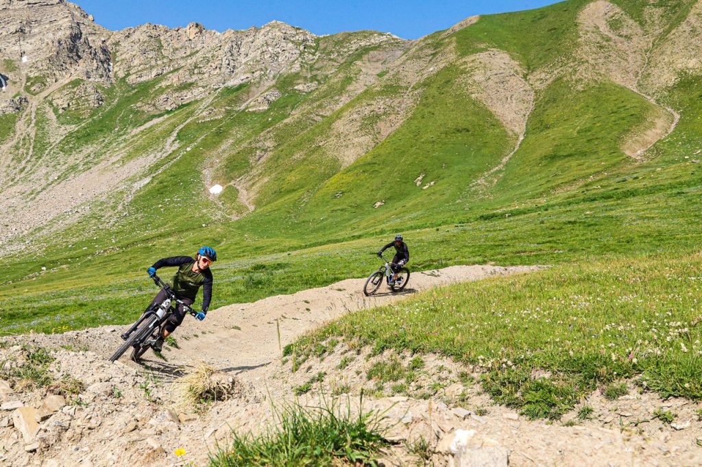 Two mountain bikers navigate a winding dirt trail through a green, rocky mountain landscape under a clear blue sky, helmets on and leaning into the turn.