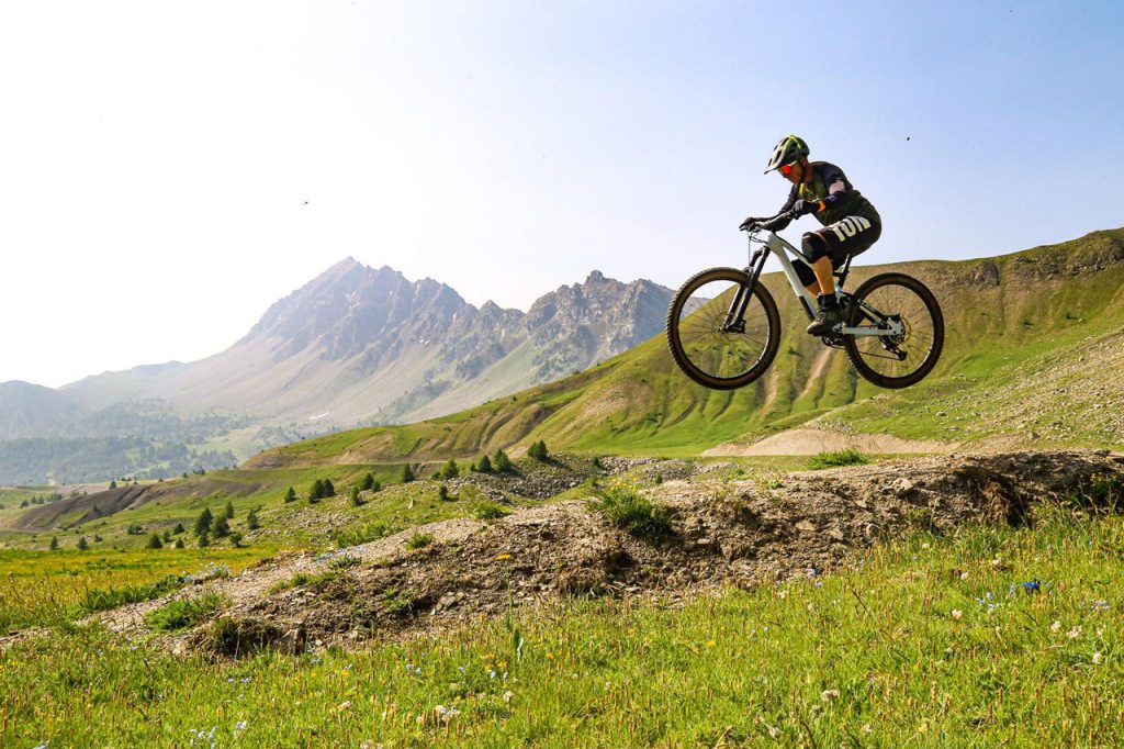 Mountain biker mid-air over a rocky trail with green hills and distant mountains in the background, under a clear sky.