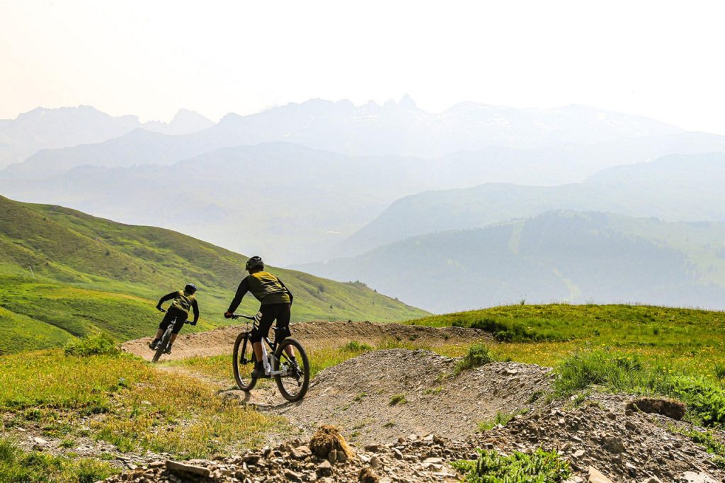 Two mountain bikers ride down a rocky dirt trail across green hills with hazy, snow-capped mountains in the distance on a clear day.