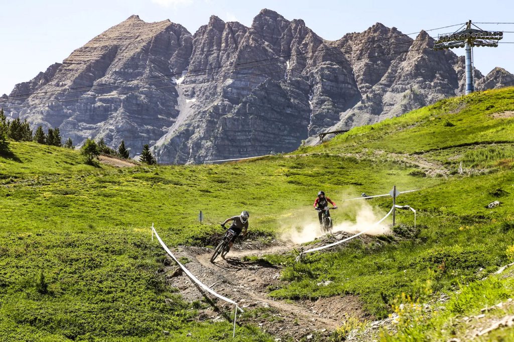 Two mountain bikers race along a dirt trail on a grassy hillside with dust clouds in the air and rugged mountains in the background.