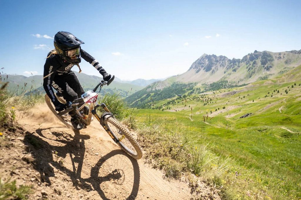 Mountain biker in full protective gear leans into a turn on a dusty hill trail with green valleys and mountains in the background