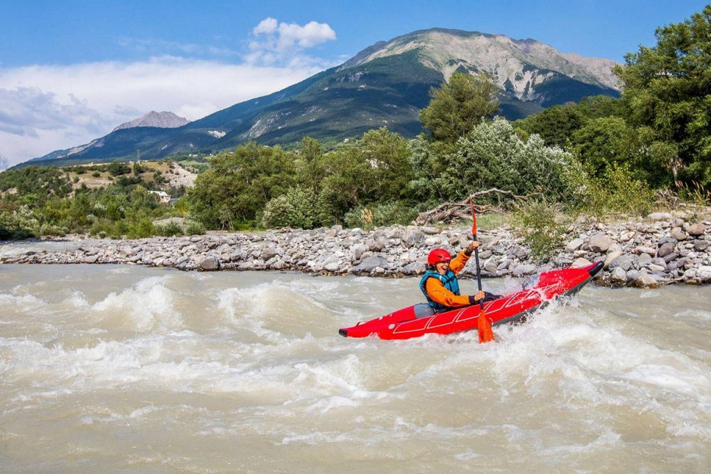 Person in a red kayak paddling through whitewater on a river with rocky bank and mountainous backdrop.