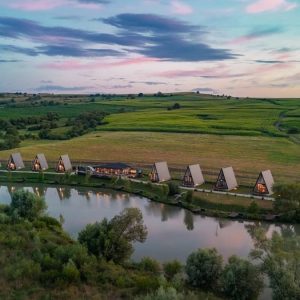 A row of A-frame cabins along a calm lake at dusk, with green fields in the background and a pink-blue sky.