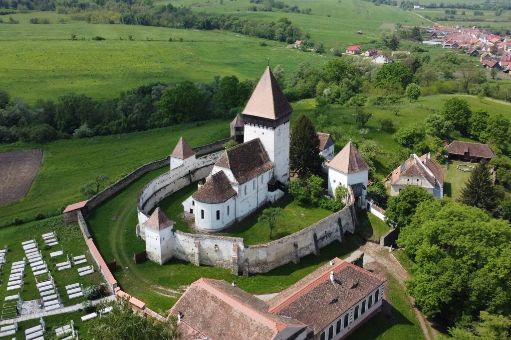 Aerial view of a white-walled castle with red-tiled roofs, central tall cone-shaped tower, surrounded by green fields and a village in the distance.