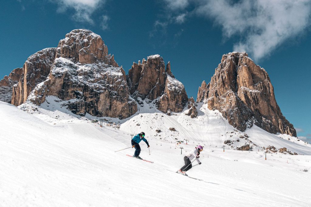 Skier in a blue jacket carving down a snowy slope with rugged mountain peaks against a clear blue sky.