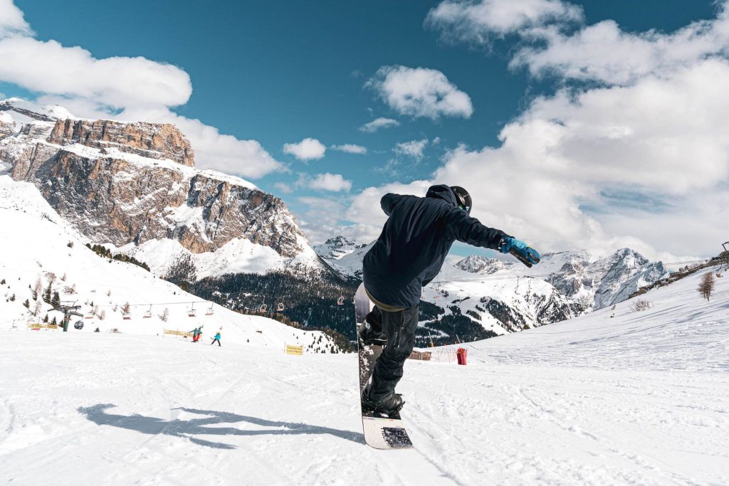 Snowboarder performing a mid-air trick on a snowy slope with rugged mountains and chairlifts in the background under a blue sky.