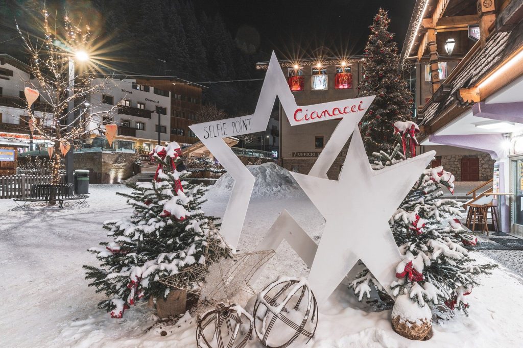 Snowy town square at night featuring a large white star sculpture labeled 'Selfie Point' and festive lights on trees and buildings.