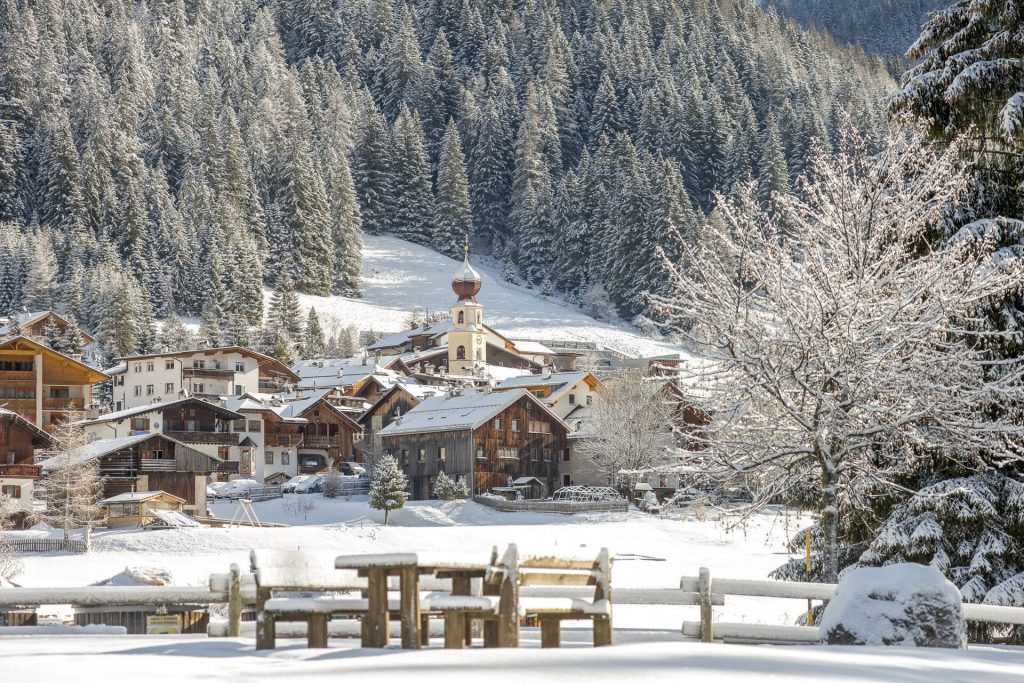 Snowy alpine village with a church steeple, wooden houses, and a forest backdrop covered in snow.