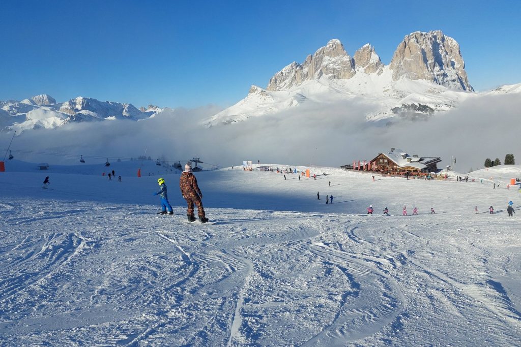 Ski slope with skiers and a wooden lodge, snow-covered mountains rise in the distance under a clear blue sky.