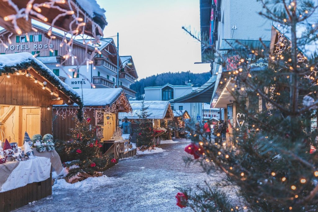 Snowy Christmas market street with wooden stalls, string lights, and shoppers near Hotel Rosa in a wintry village.