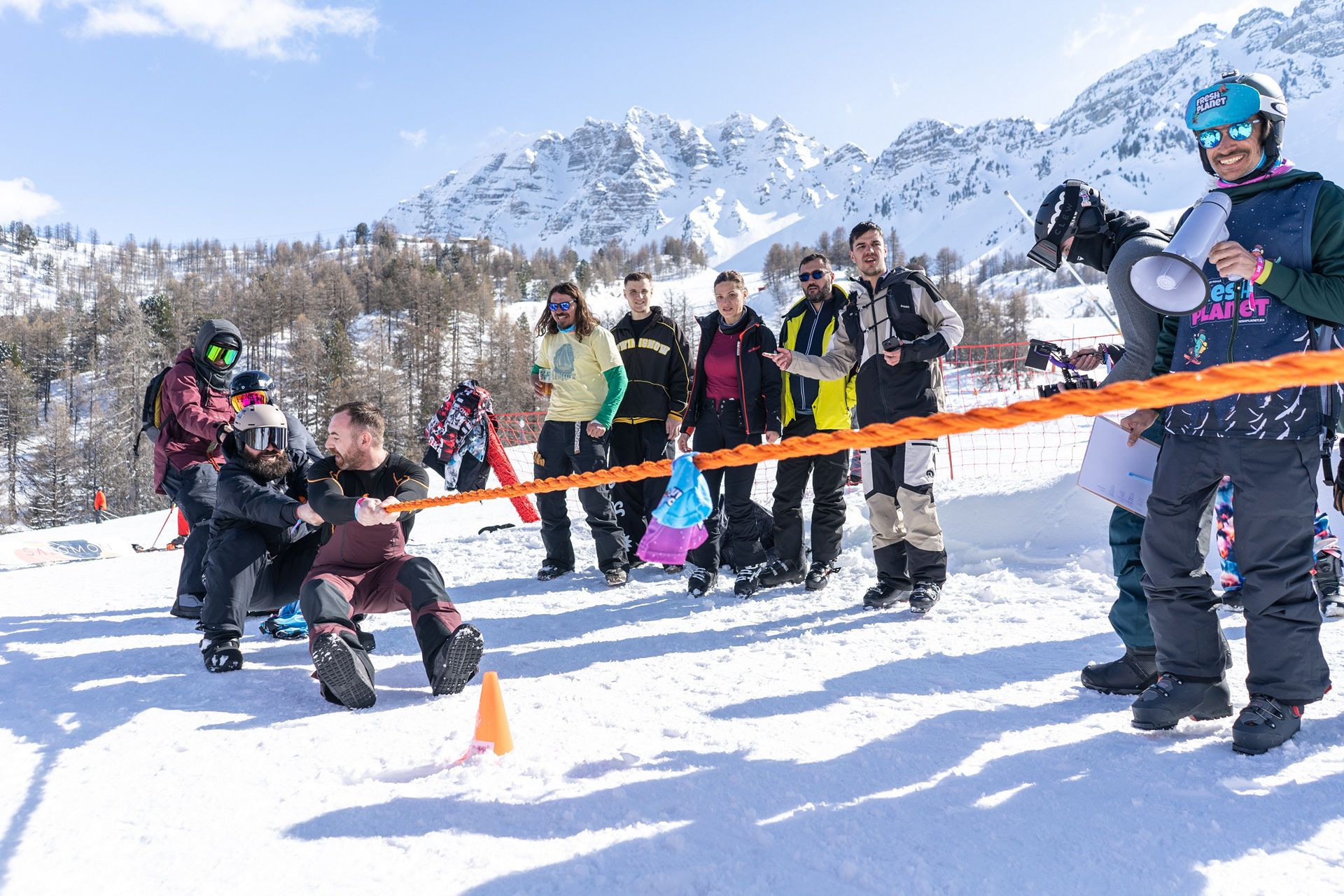 ADR05113 snow vars | Fresh Planet | Fresh Planet Group of skiers and snowboarders in winter gear watching a rope-pulling game on a snowy slope, with snow-capped mountains in the background