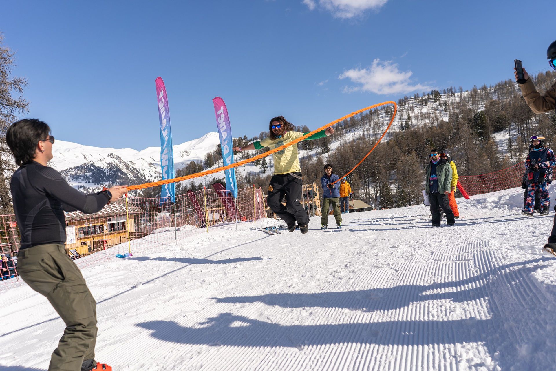 ADR05324 snow vars | Fresh Planet | Fresh Planet Person in mid-air jumping with an orange rope in a snowy slope, others watching in winter gear against a mountain backdrop with blue sky.