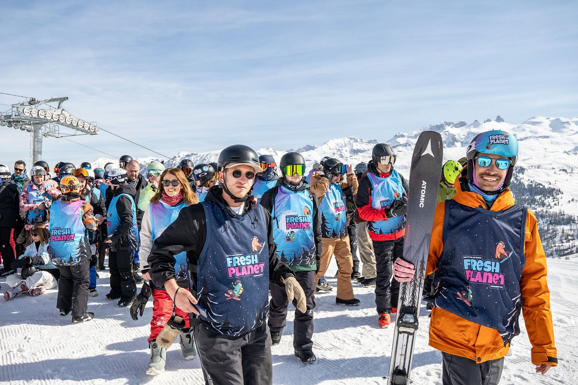 Crowd of skiers in blue'Fresh Planet' vests on a snowy slope with mountains in the background.