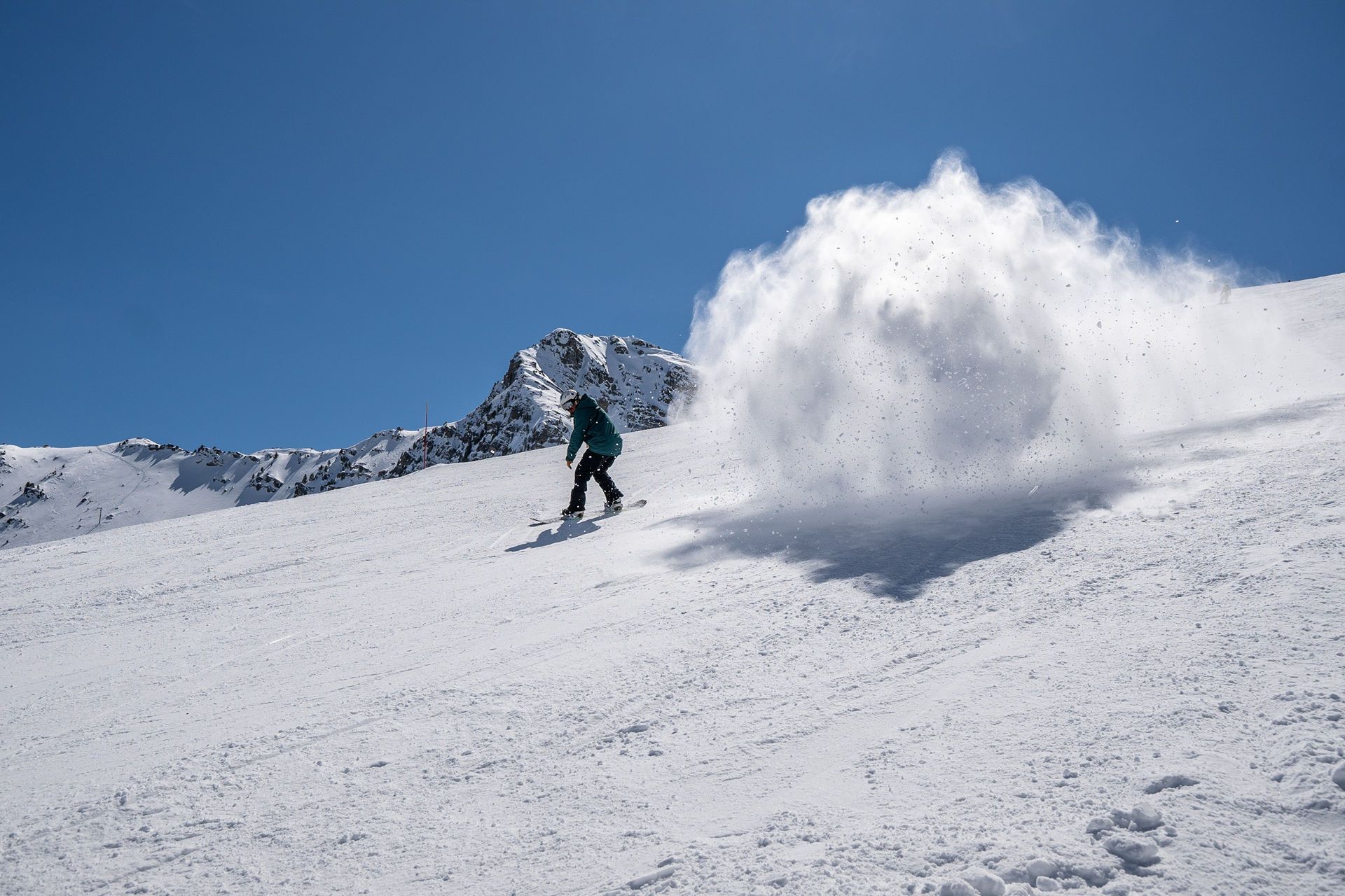 Snowboarder on a snowy slope kicking up a large cloud of snow against a clear blue sky.
