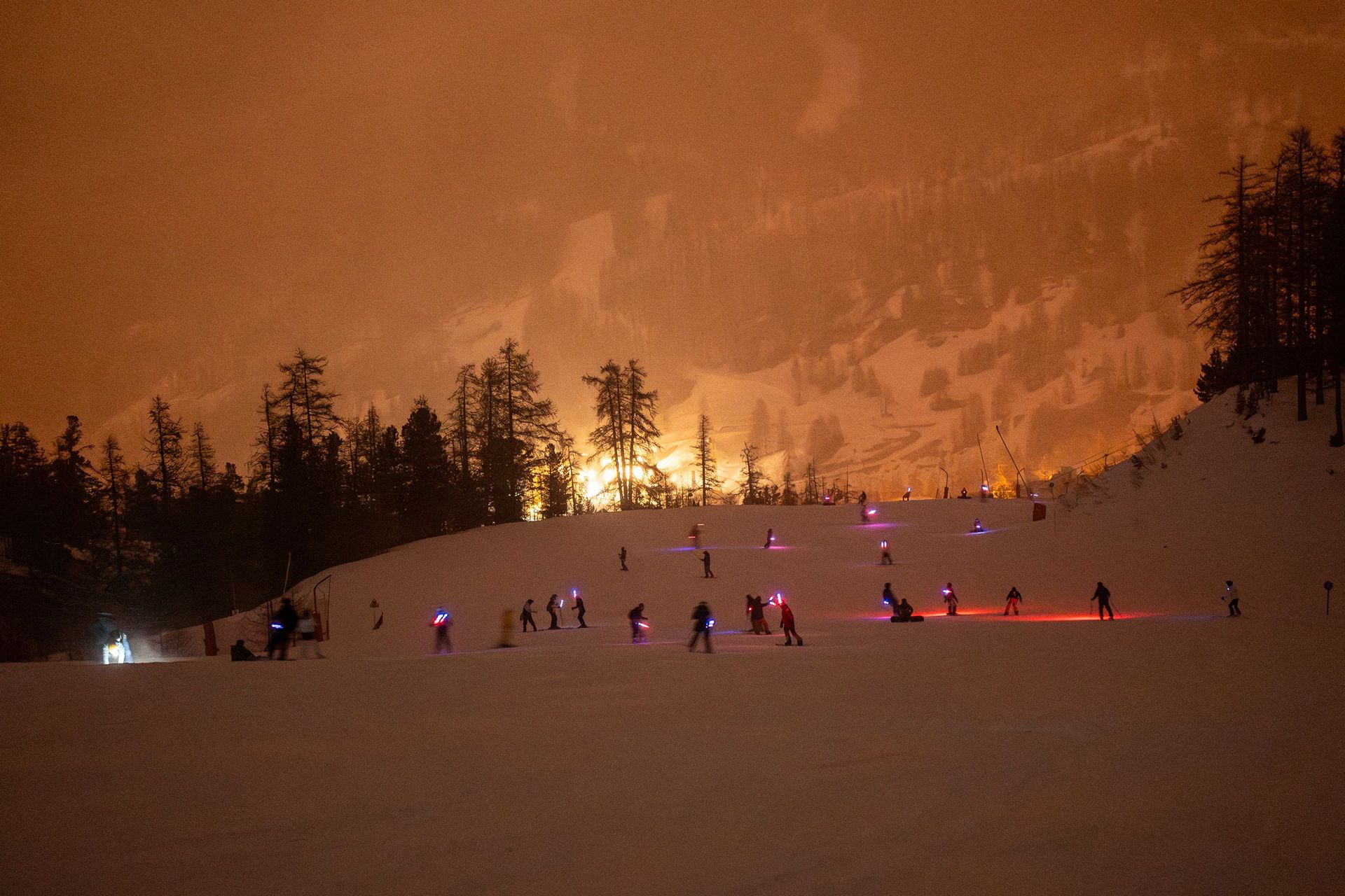 Night ski scene with many skiers illuminated by colorful lights on a snow slope, orange glow lighting the mountainside and trees in the background.