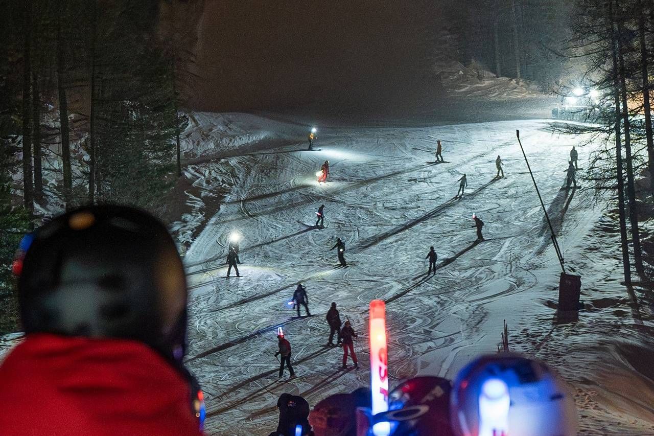 Nighttime ski slope with illuminated skiers and foreground helmets; a group glides down under headlamps.