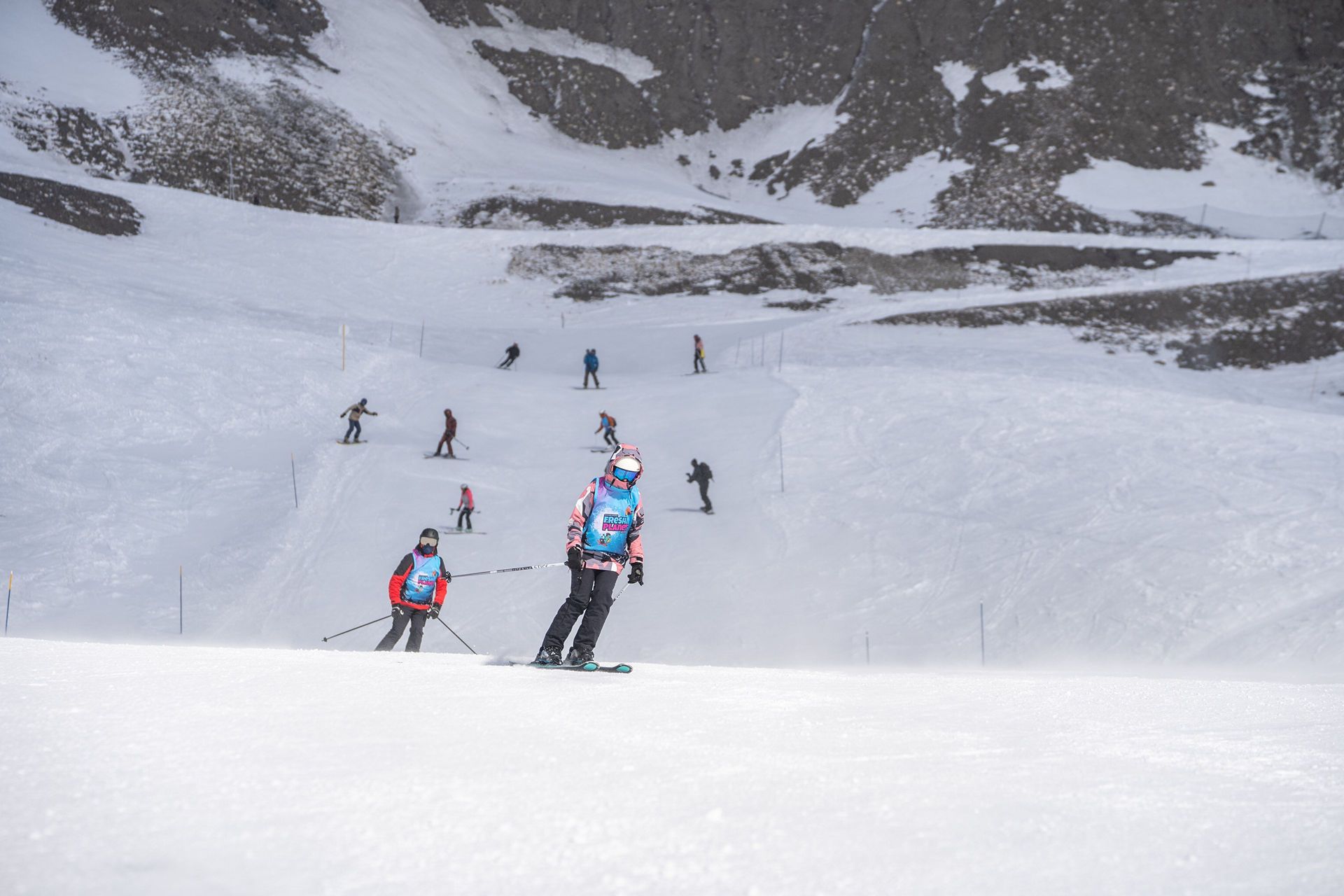 ADR09054 snow vars | Fresh Planet | Fresh Planet Snowy ski slope with multiple skiers, two in bright blue jackets leading in the foreground, and rocky cliffs in the background.