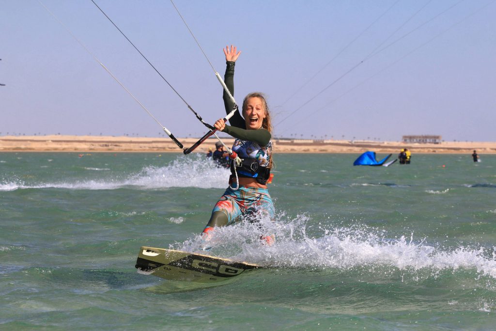Kiteboarder with colorful wetsuit skating on turquoise water, raising left hand in the air as spray splashes around. In the background, a sandy shore and other kiters are visible under a clear blue sky.