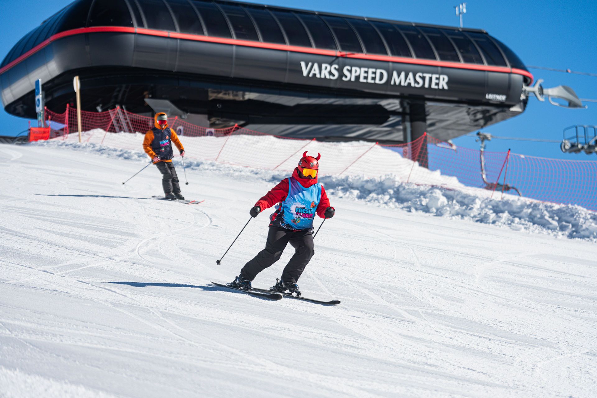 Two skiers descend a snowy slope with a large black ski lift station behind them reading'VARS SPEED MASTER' on a clear blue sky.