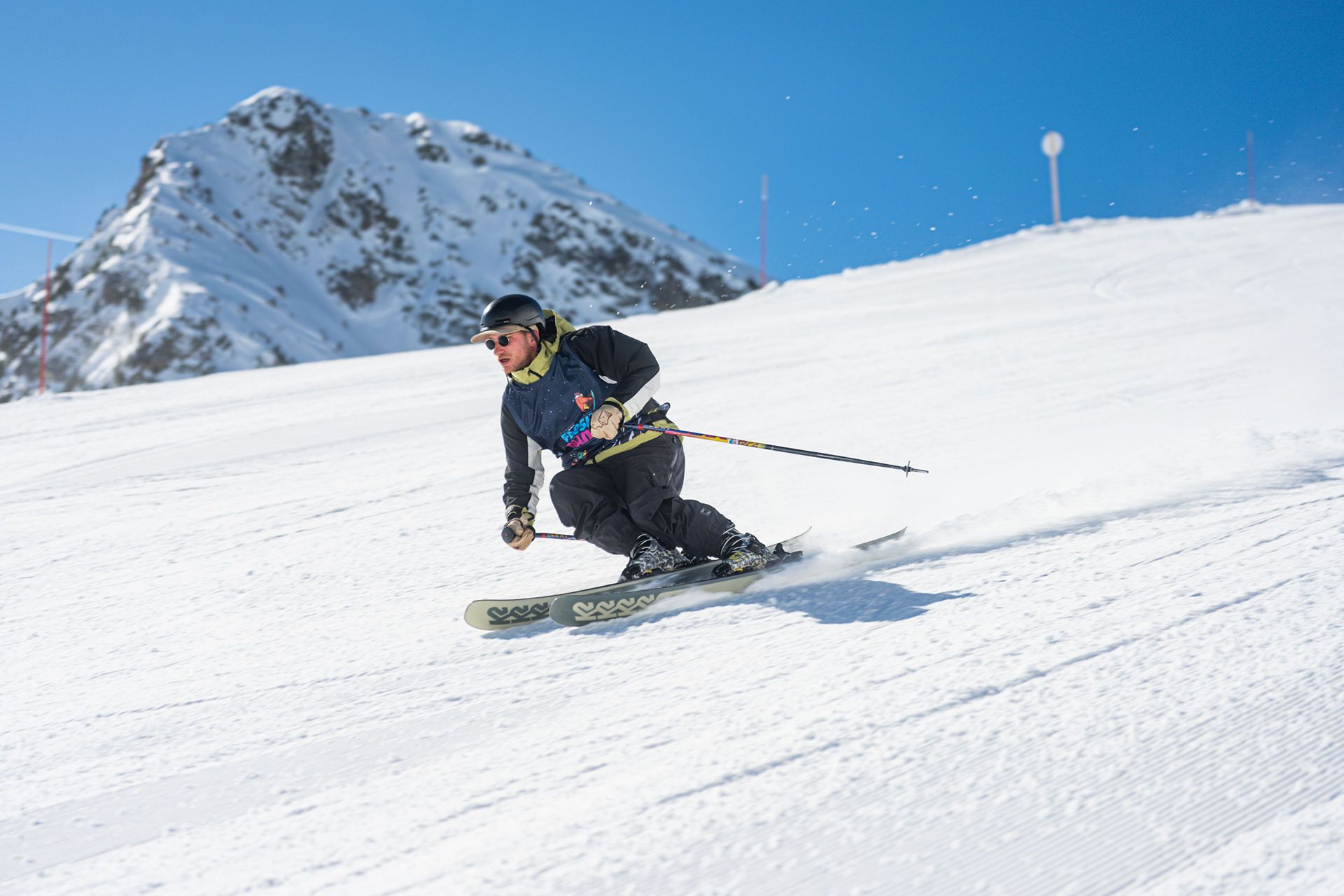 BLJ04478 snow vars | Fresh Planet | Fresh Planet Skier wearing helmet and goggles making a sharp turn on a sunny, groomed slope with a snow-covered mountain in the background